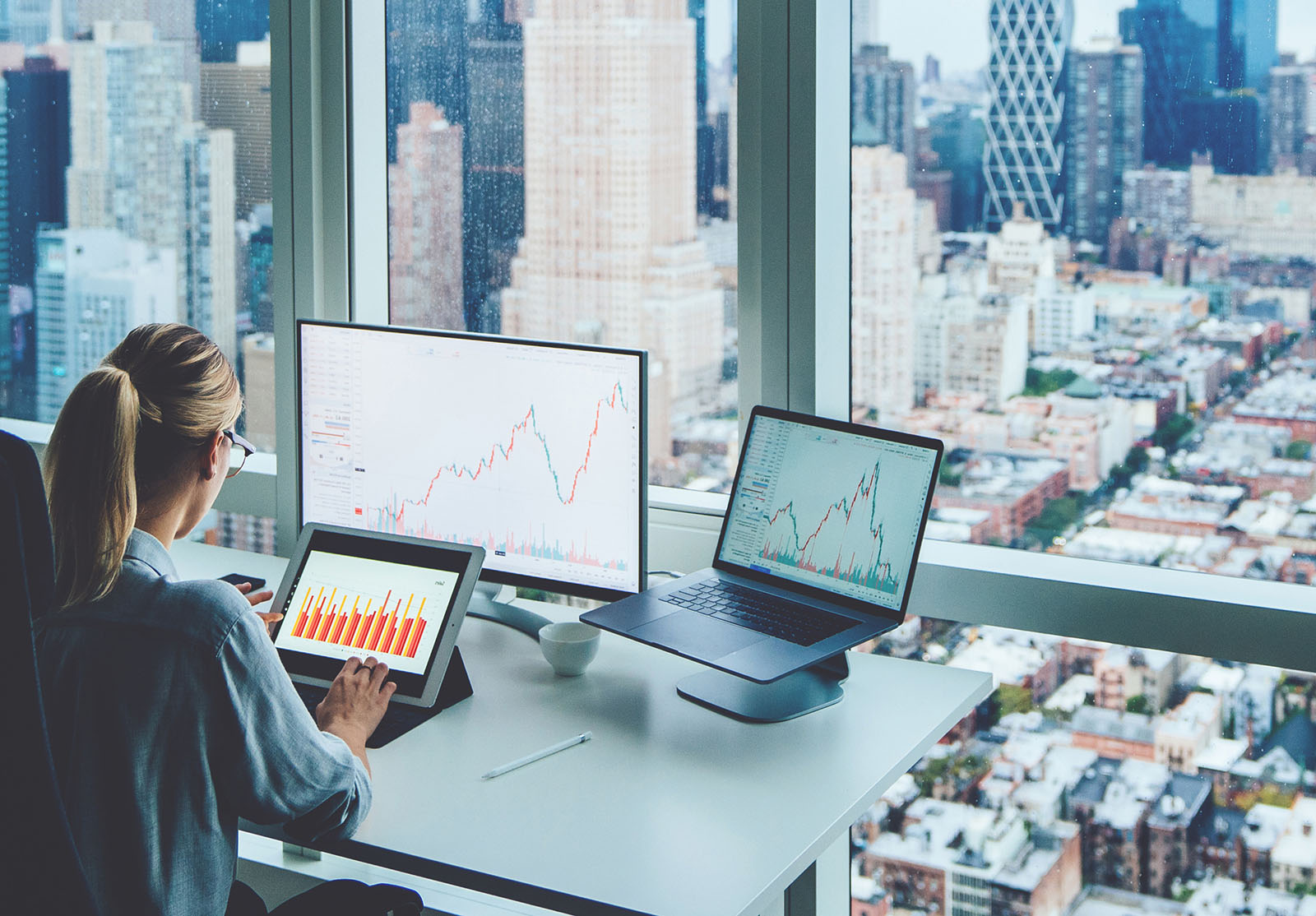 Vue de dos d'une femme d'affaires assise au bureau panoramique d'un gratte-ciel devant un ordinateur PC avec des graphiques financiers et des statistiques sur le moniteur.Analyse du marché numérique et de l'investissement dans la chaîne de blocs.