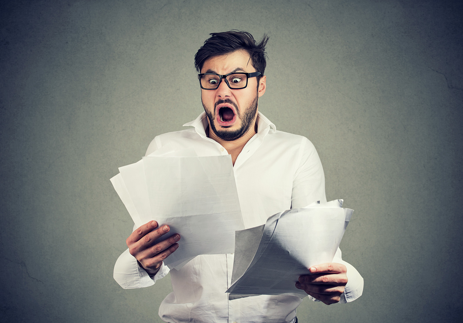 Jeune homme choqué, portant des lunettes, regardant des documents bancaires avec une liste de factures et de frais et se sentant stressé sur fond gris.