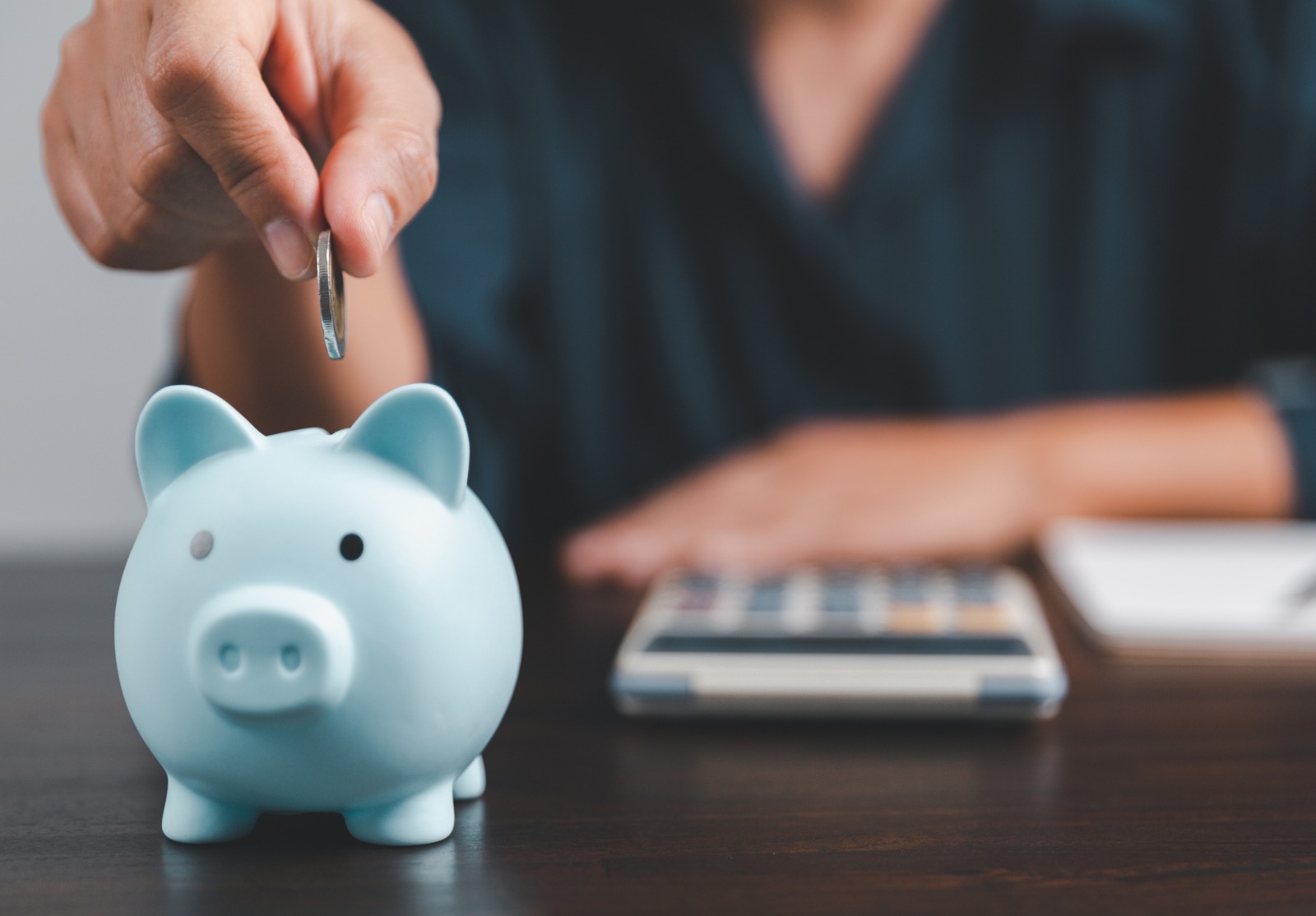 Une personne assise derrière une calculatrice et prête à mettre une pièce dans une tirelire bleue.