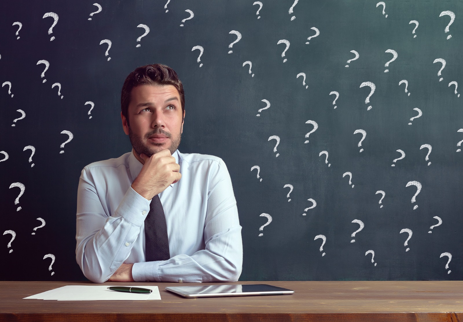 Homme d'affaire qui semble réfléchir assis à une table. On voit plein de points d'interrogation flotter autour de lui.