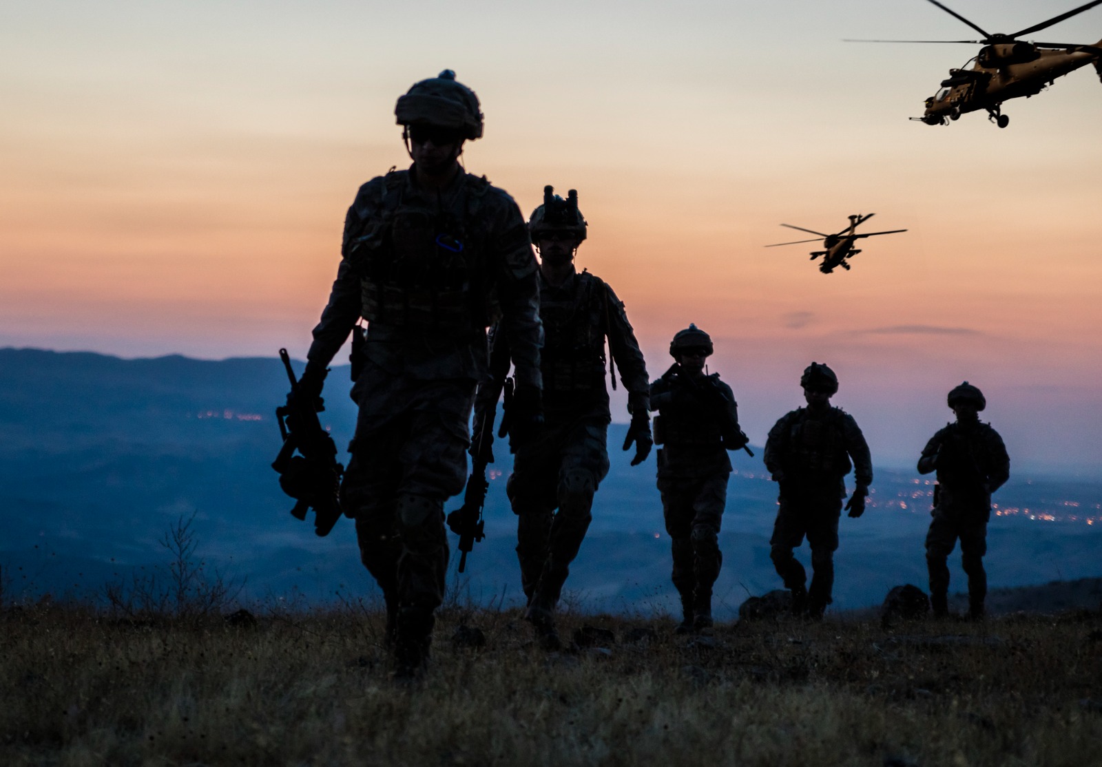 Plusieurs militaires marchant dans une plaine au crépuscule. Dans le ciel, on voit deux hélicoptères.