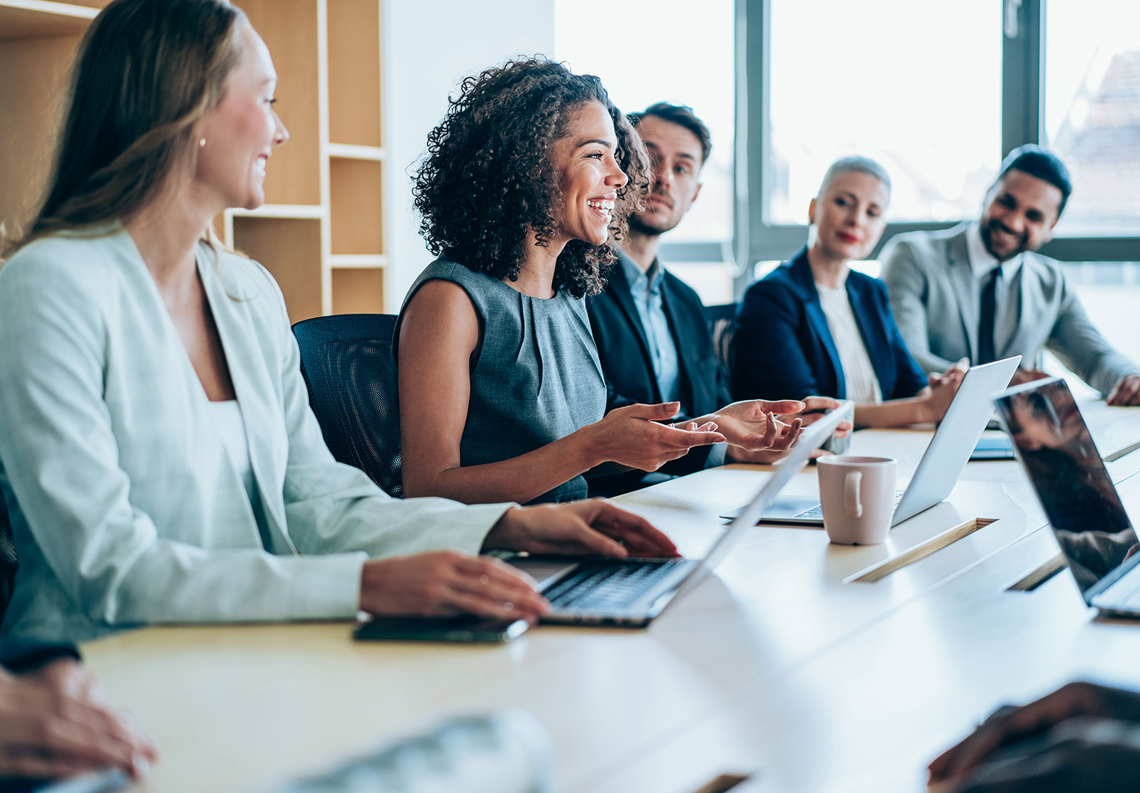 Des hommes et des femmes d'affaire assis à une table écoutent une femme en robe qui sourit et qui semble parler.