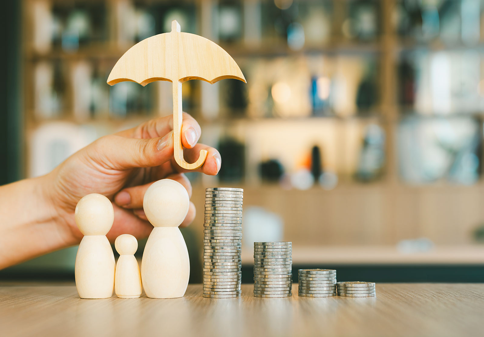 Une main tenant une forme de parapluie en bois au-dessus de trois formes représentant des parents et un enfant et de piles de monnaie.