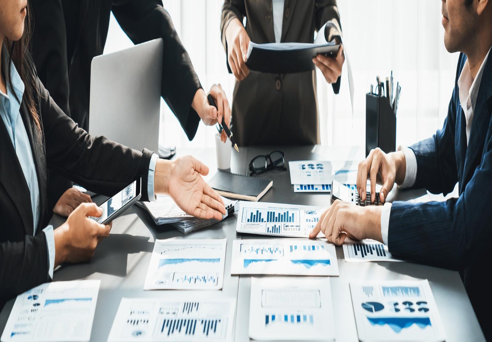Groupe de professionnels en réunion, entourés de nombreux documents financiers et graphiques, en pleine discussion.