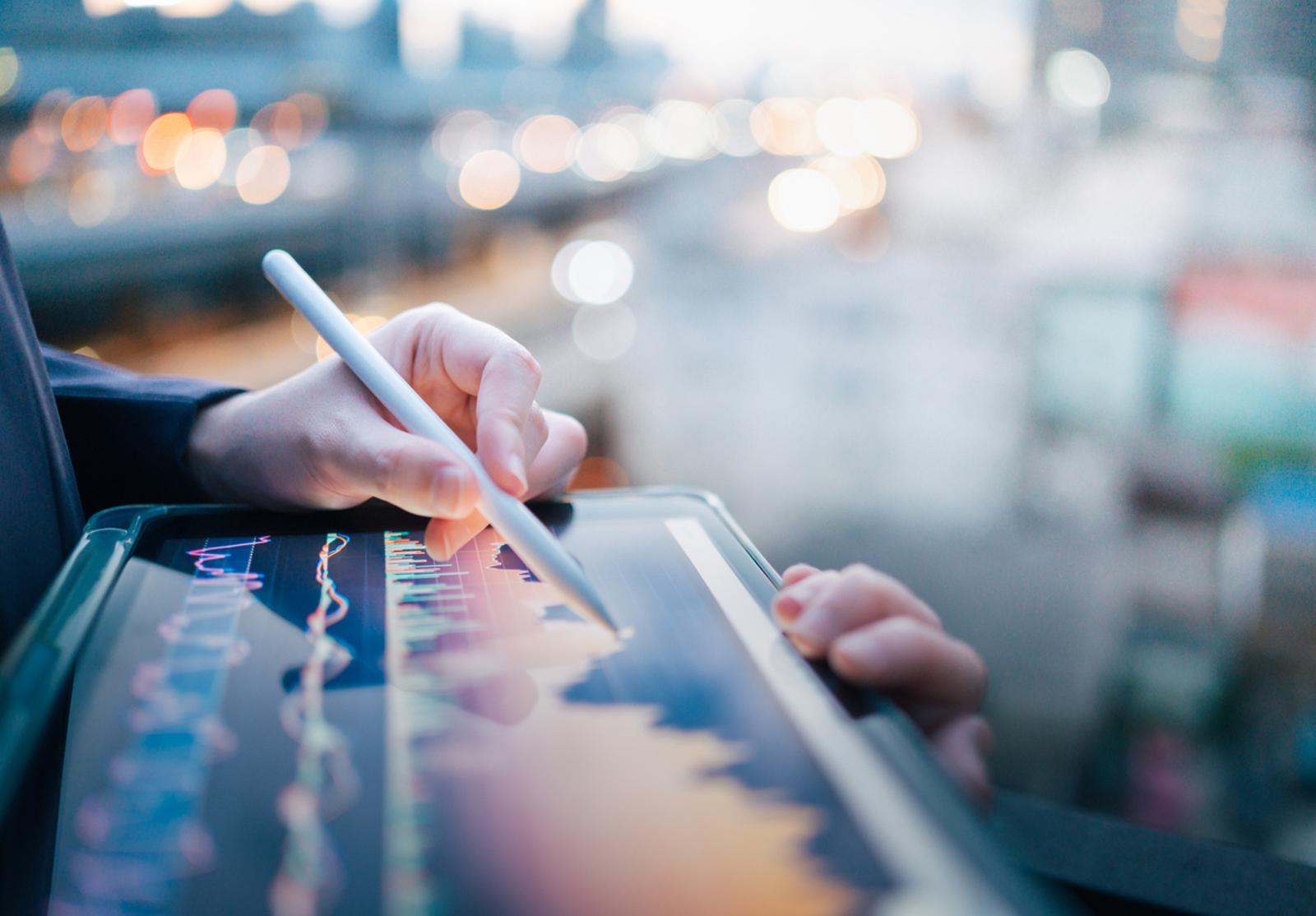 Main tenant un stylet au-dessus d’une tablette affichant un graphique boursier interactif, en contexte urbain flou.