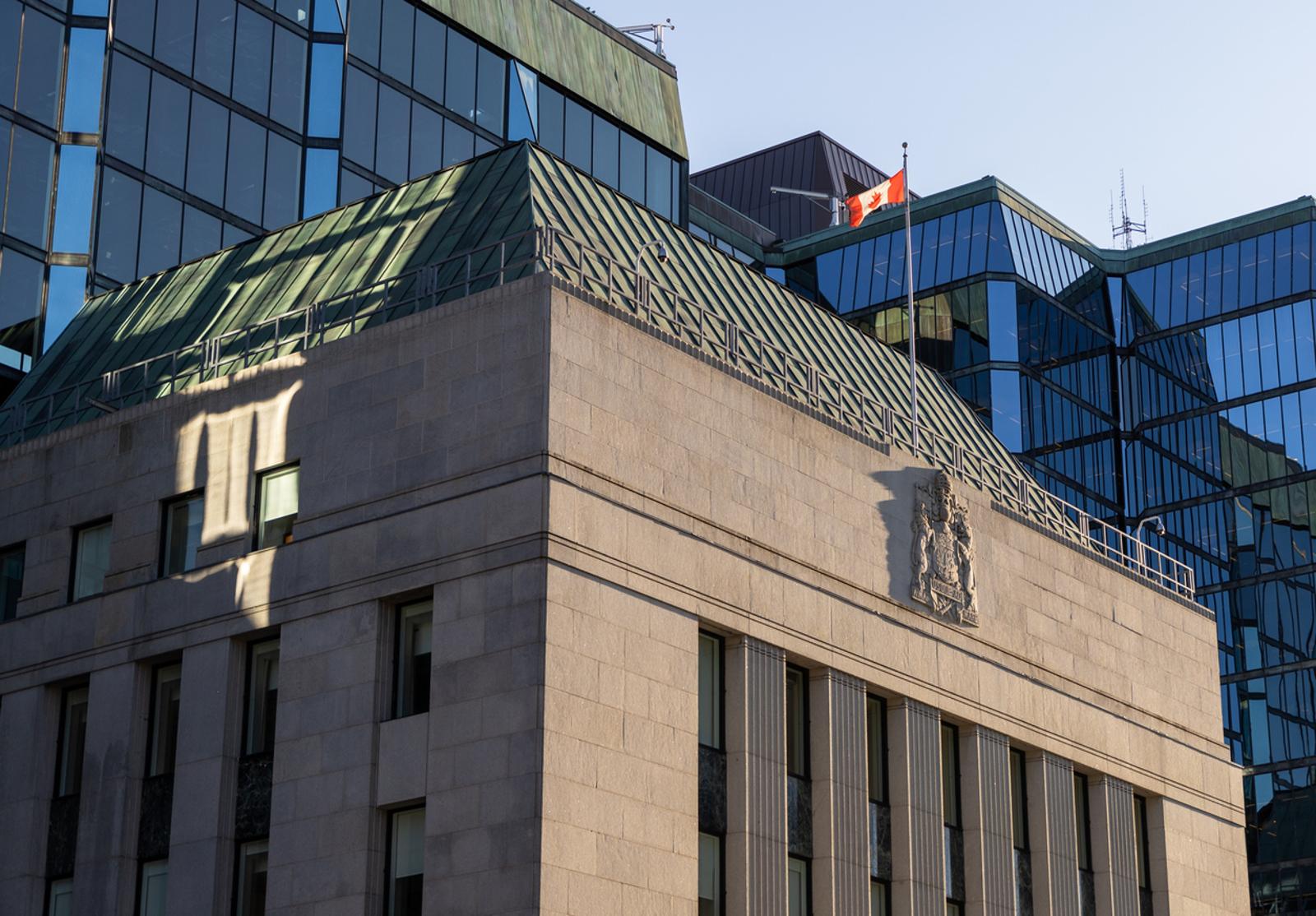 Façade du bâtiment moderne de la Banque du Canada, avec un toit vert, le drapeau canadien et des reflets sur les vitres.