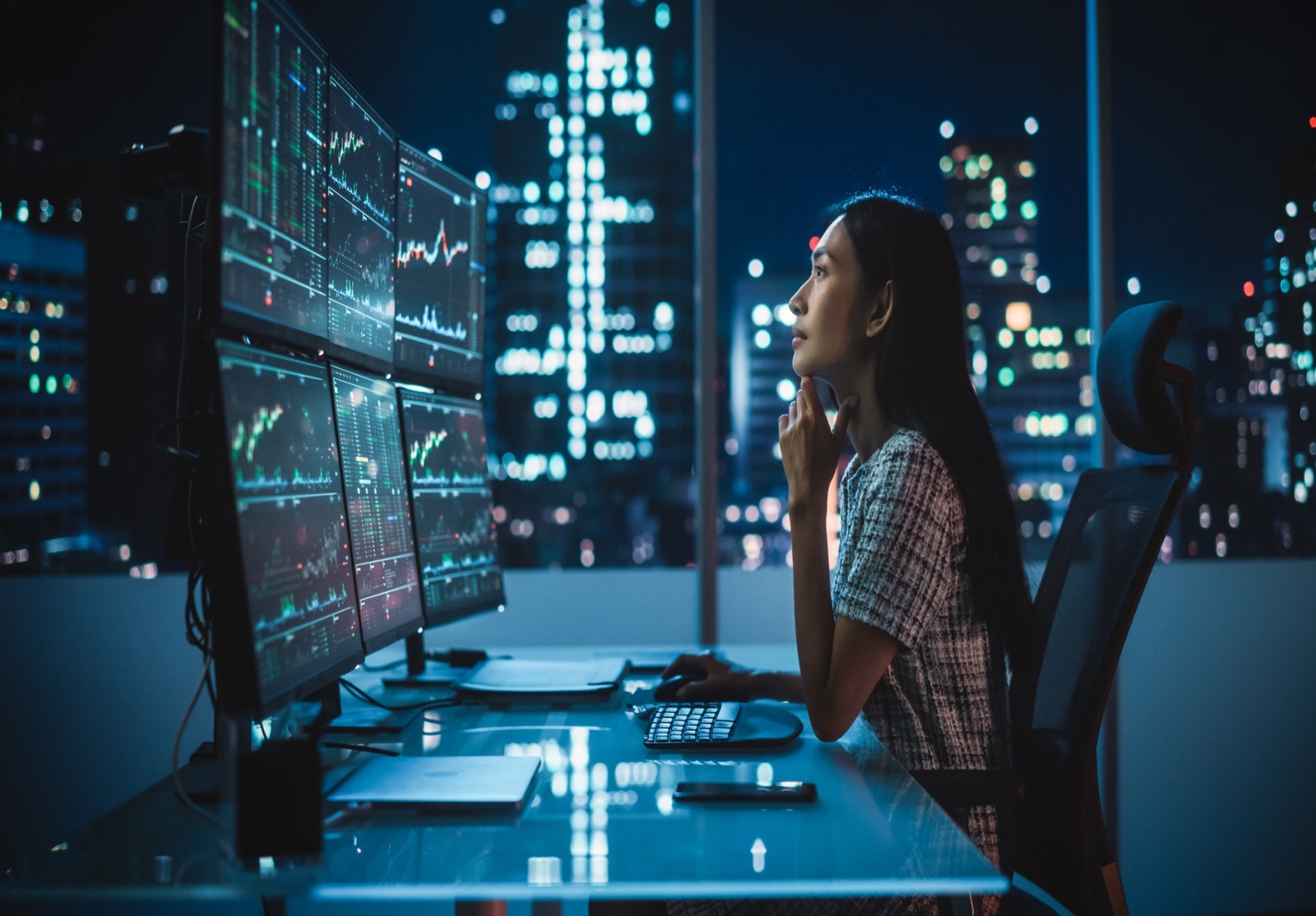 Une professionnelle travaillant tard dans la nuit dans un bureau moderne avec vue sur la ville illuminée. Elle est assise devant plusieurs écrans affichant des graphiques financiers et des données boursières en temps réel, illustrant l'analyse des marchés financiers, le trading et la technologie.